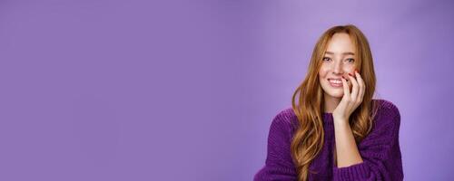 Close-up shot of feminine and glamour cute redhead girl with freckles in purple knitted sweater holding hand on cheek gently chuckling and smiling broadly at camera over violet background photo
