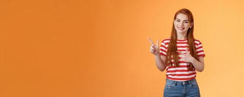 Cheerful pleasant female professional assistant showing customers office, pointing left smiling cheerful, gladly help out pick product, introduce new promos copy space, stand orange background photo