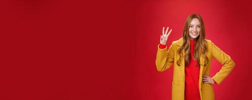 Cute friendly-looking ginger girl showing three reasons buy product smiling broadly making third reservation, looking delighted and confident in her choice posing against red background in yellow coat photo