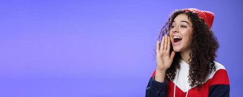 Friend shouting through street to turn attention. Portrait of carefree happy cute woman in beanie and sweatshirt holding palm near opened mouth looking with smile left as calling mate with loud yell photo