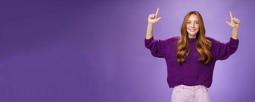 Scroll up and see cool copy space. Portrait of happy attractive and friendly young ginger woman with freckles in warm purple sweater raising hands pointing upwards as showing place photo
