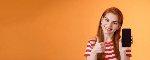 Best smartphone app guarantee. Assertive lovely friendly-looking redhead woman recommend cellphone application, introduce online store website, like give thumb-up approval, stand orange background photo