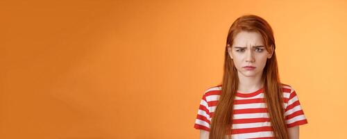 Close-up moody upset childish redhead girl, sulking unfair situation, frowning upset, complaining disappointed, pouting offended, stand uneasy perplexed orange background, whining displeased photo