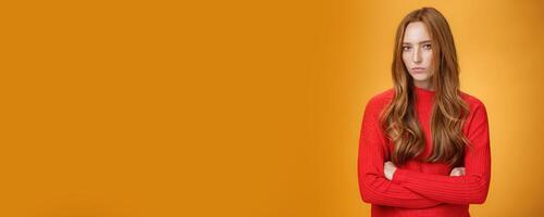 Suspicious intense and defensive ginger girl standing in passive-aggressive pose pouting and frowning looking with disbelief and disdain at camera, offended against orange background photo