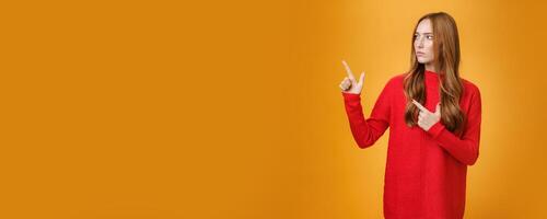 Studio shot of suspicious serious-looking unsure redhead girl frowning looking and pointing at upper left corner with doubtful strict gaze posing uncertain over orange background photo
