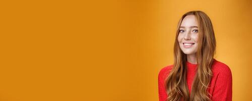 Close-up shot of happy charismatic and carefree cute redhead girl in red knittes outfit smiling joyfully as turning at camera satisfied and joyful posing delighted over orange background photo