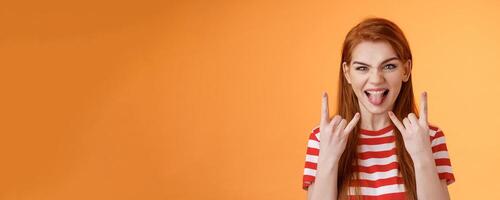 Lets rock-n-roll. Daring cheerful good-looking caucasian redhead woman, having fun, show heavy metal gestures, stick tongue and smile amused, enjoy music festival, stand orange background photo