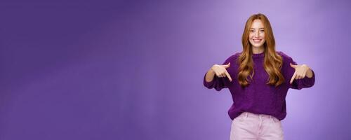 Girl giving recommendation to visit place, pointing down and smiling friendly with delighted and pleasant grin. Portrait of attractive redhead female model in purple warm sweater promoting copy space photo