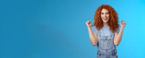 Hooray successful day. Cheerful chrismastic redhead girl summer dungarees clench fists joyfully smiling broadly determined achieve success, triumphing winning game standing upbeat blue background photo