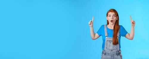 Studio shot amazed curious redhead woman drop jaw astonished, look pointing ambushed top copy space, check out up advertisement thrilled, stand surprised blue background open mouth photo
