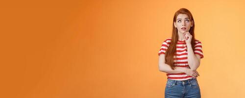 Thoughtful cute curious redhead woman looking intrigued up, touch chin thinking, making assumption, observe interesting copy space product, stand orange background pondering information photo