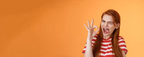 Sassy asserive relaxed redhead woman assuring everything under control, make deal, wink show tongue silly accepting terms, judgemental approval gesture, like idea, show okay excellent sign photo