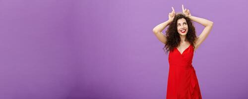 Woman being daredevil on party standing sensual and seductive in red elegant evening dress holding index fingers on head like devil horns looking at upper right corner with flirty smile photo
