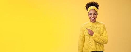 Friendly good-looking enthusiastic black charming girl in headband sweater smiling broadly amused pointing left awesome place showing you perfect blank space advertisement, posing yellow background photo