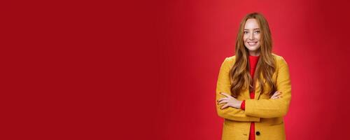 Charming successful young female in yellow autumn coat smiling broadly holding hands crossed on body in confident self-assured pose standing delighted and happy over red background photo