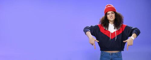 Snobbish and displeased stylish and cool street girl with curly hair in beanie and sweatshirt pointing down grimacing as feeling unsatisfied and disappointed posing against blue background photo
