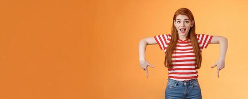 Enthusiastic lively cute redhead woman introduce incredible offer, pointing down, bottom copy space, smiling amazed, look camera, explain you awesome opportunity, feel excitement, orange background photo