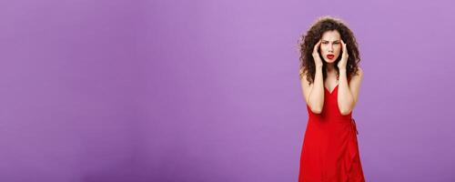 Stupid people getting on her nerves. Irritated and bothered stylish rish businesswoman in evening red dress pulling corners of eyes aside staring annoyed and unfocused at camera over purple background photo