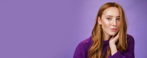 Sensual and romantic woman with red hair and freckles touching cheek gently as curls dropping on half face gazing charmed and flirty at camera, smiling being tender and coquettish over purple wall photo