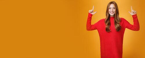Enthusiastic and excited energized redhead female in red warm sweater pointing up with rised hands and smiling broadly thrilled with awesome promotion, feeling astonished against orange background photo