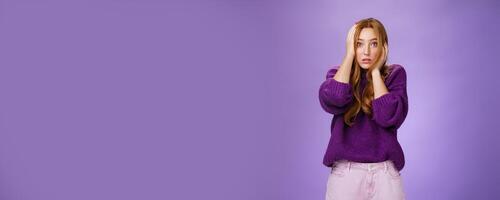 Girl in stupor feeling shocked and troubled as realising huge mistake made, holding hands on face and head popping eyes at camera confused and hopeless, posing anxious over purple background photo