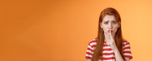 Uneasy worried insecure redhead girl begging keep secret safe, frowning grimace upset, shushing, press index finger lips hush gesture, worry someone spread rumor, orange background photo