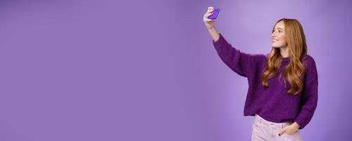 Attractive confident and stylish happy 20s woman with ginger hair standing carefree with joyful smile over purple background extending hand with smartphone as taking selfie happily photo
