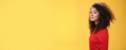 Profile shot of stylish and cool attractive curly-haired european 25s woman in red dress turning at camera and smiling broadly with delighted carefree expression, having fun over yellow background photo