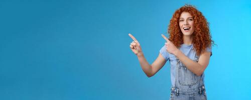 Hey check this out. Enthusiastic redhead curly stylish summer girl inviting beach party pointing upper left corner directing awesome location smiling broadly discuss awesome promo blue background photo