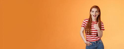 Enthusiastic amazed sassy modern redhead female attend impressive bad awesome design, look amused, observe cool place, pointing left excited, open mouth fascinated intrigued, stand orange background photo