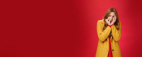 Portrait of shocked and scared insecure woman in yellow coat covering face in horror and fear biting nails squinting and looking intense left as frightened being victim standing in shook, trembling photo