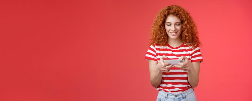 Redhead girl fool around waiting queue dentist playing awesome smartphone game hold phone horizontal tap cellphone screen look telephone display smiling delighted entertained standing red background photo
