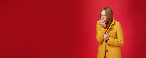 Girl getting cold, coughing and buttoning yelling light coat as freezing on chilly, windy weather looking displeased and ill at camera, feeling unwell catching flu over red background photo