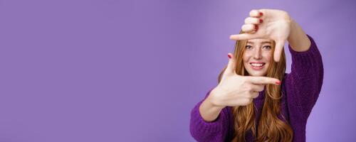 Imagine if. Portrait of hopeful and carefree creative charming redhead 20s woman showing frame with pulled hands and smiling through it as picturing bright and happy future over purple background photo