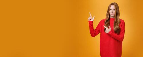 Intense suspicious and doubtful european ginger girl in warm sweater frowning hesitant and confused pointing at upper left corner asking question, looking with disbelief over orange background photo