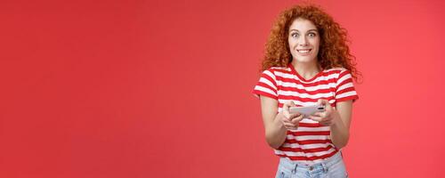 Excited happy cute redhead curly-haired girlfriend eager start learn how play boyfriend game look amused cheerful camera smiling joyful hold horizontal smartphone load app red background photo