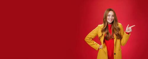 Stylish and self-assured attractive redhead woman with freckles in yellow outdoor coat pointing right with finger pistol and smiling joyfully as camera as showing cool promotion over red wall photo