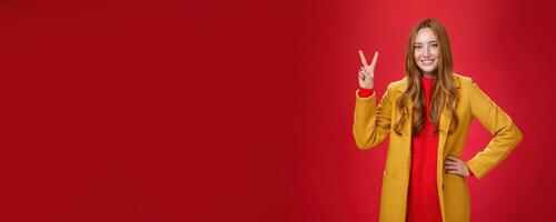 Portrait of happy playful and cute redhead woman with freckles in yellow coat holding hand on waist confident and self-assured showing peace or two sign delighted, beating weather conditions photo