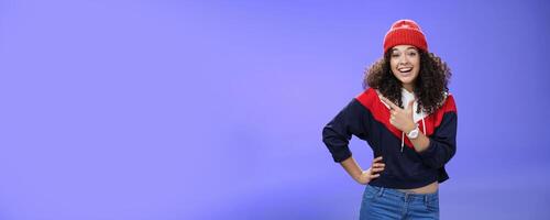 Stylish and sociable friendly-looking attractive woman with curly hair in warm beanie pointing at upper left corner laughing from joy and amazement having fun posing happily over blue background photo