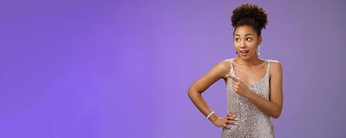 Curious unsure african-american attractive elegant young woman in silver glittering evening dress look pointing left intrigued discuss strange person gaze doubtful uncertain, blue background photo