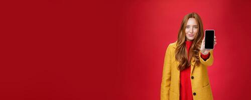 Attractive and stylish ginger girl with confident glance and self-assured smile pulling mobile phone at camera showing smartphone screen grinning delighted as posing against red background photo