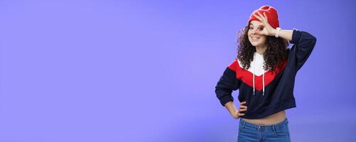 Okay girl agrees. Friendly-looking happy and charming young curly-haired girl in warm beanie showing ok gesture or circle over eye as peeking at camera and smiling broadly with joy over blue wall photo