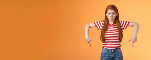 Cute timid hesitant ginger female taking tough decision feel pressured upset, frowning, pulling gloomy unhappy face, pointing down disappointed, uneasy taking decision, orange background photo