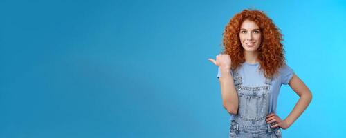 Cheerful curious lively redhead good-looking curly-haired girl wearing summer overalls t-shirt pointing left thumb look camera intrigued excited explore new stores friends walking shopping mall photo