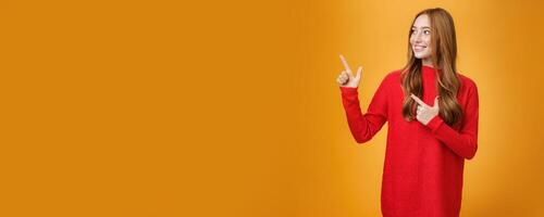 Interested gentle and tender young 20s ginger woman in red warm sweater pointing, looking at upper left corner with broad smile, enjoying nice happy scene over orange background photo