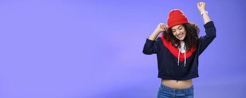 Girl celebrating favorite season in year. Portrait of carefree and joyful dancing woman with curly hair in cute red hat lifting hands in dance movements smiling enjoying music and holidays photo