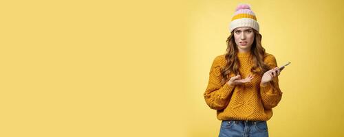 Confused cringing annoyed young woman shocked how friend talked her frowning raise hand dismay look frustrated camera holding smartphone finish call unpleasant note, yellow background photo