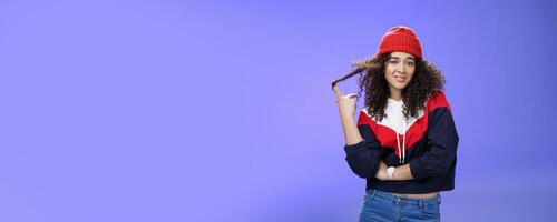Stylish and snobbish arrogant curly-haired woman in warm beanie rolling curl on finger smirking and looking with contempt at camera, scorning person as being too cool over blue background photo