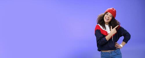 Studio shot of enthusiastic charming curly-haired woman inviting to play in snow with her pointing at upper right corner tilting head joyfully and smiling, holding hand on waist over blue wall photo