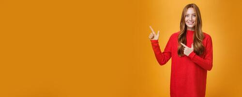 Assertive and helpful charming young female assitant in red knitted dress with ginger hair smiling friendly and pleasant as pointing at upper left corner, showing interesting thing over orange wall photo